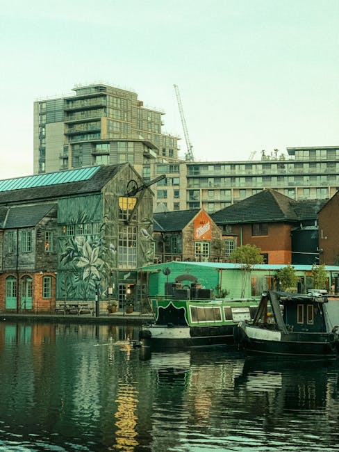 A view of a residential and commercial area near Putney Wharf, featuring a canal with two boats moored along the edge. In the foreground, the water on the canal appears calm and reflects the surrounding buildings. On the left, there is a rustic, weathered building with greenish wooden siding and a mural of tropical foliage painted on its side. Adjacent to it, a modern brick building with multiple windows and a sloped roof is visible. In the background, a multi-storey urban development with glass balconies, concrete structures, and a construction crane indicates ongoing development. Natural daylight illuminates the scene, highlighting the clean surfaces of the boats and the well-maintained appearance of the buildings. Putney Carpet Cleaning's professional services could assist in maintaining the hygiene of similar residential surfaces and properties in the area, ensuring a high standard of cleanliness and surface sanitisation in apartments along Putney Wharf.