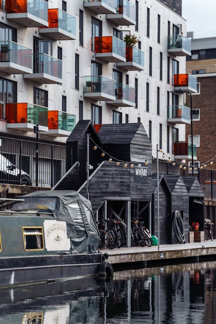 A view of a residential and commercial area near Putney Wharf, featuring a canal with two boats moored along the edge. In the foreground, the water on the canal appears calm and reflects the surrounding buildings. On the left, there is a rustic, weathered building with greenish wooden siding and a mural of tropical foliage painted on its side. Adjacent to it, a modern brick building with multiple windows and a sloped roof is visible. In the background, a multi-storey urban development with glass balconies, concrete structures, and a construction crane indicates ongoing development. Natural daylight illuminates the scene, highlighting the clean surfaces of the boats and the well-maintained appearance of the buildings. Putney Carpet Cleaning's professional services could assist in maintaining the hygiene of similar residential surfaces and properties in the area, ensuring a high standard of cleanliness and surface sanitisation in apartments along Putney Wharf.