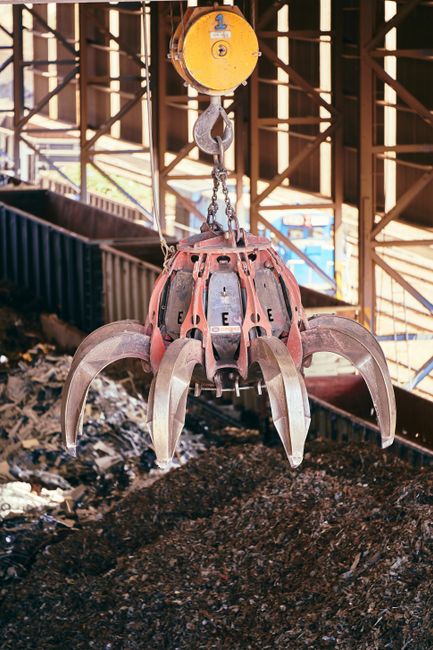 A large industrial claw attachment hanging from a yellow overhead crane, positioned above a pile of shredded textile waste on a concrete floor within an industrial facility. The claw is made of metal with a reddish hue, featuring multiple curved tines and labeled with the letter 'E.' The background reveals a structure of steel beams and wooden panels, with natural light filtering through openings, highlighting the cleanliness of the surrounding surfaces and equipment. The scene focuses on materials used in heavy-duty industrial waste handling, relevant to the topic of managing bulky textile waste, and emphasizes the importance of proper handling and cleaning in textile waste disposal, offered by Putney Carpet Cleaning.