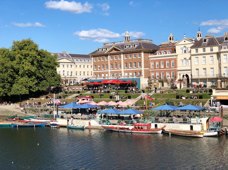 A scenic view of the Lower Richmond Road area with a promenade along the river, featuring several small boats and yachts moored at the water's edge with vibrant blue, pink, and red canopies over boat seating areas. Behind the boats, there are tiered grassy slopes with numerous visitors sitting on benches and grassy patches, enjoying the sunny weather. On the upper level, ornate multi-story buildings with classical architectural features and a mixture of brick and cream-colored facades line the street, with some having small turrets and detailed windows. The scene is brightly lit with clear blue skies and a few scattered clouds, indicating a pleasant day, and lush green trees add to the tranquil, picturesque atmosphere. This image exemplifies urban leisure spaces that can benefit from regular surface cleaning and maintenance by [COMPANY_NAME] and local cleaning services to ensure hygiene and aesthetic appeal.