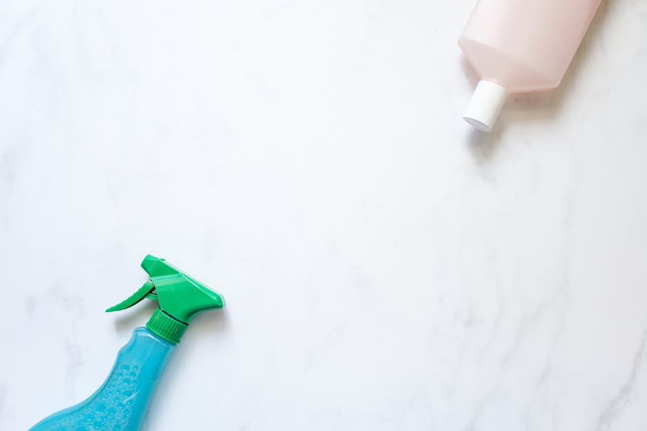 A close-up view of a cleaning spray bottle with a green trigger and blue body, and a partially visible pink bottle in the upper right corner, both placed on a clean, white marble surface. The surface is smooth, glossy, and free of dust or stains, indicating it has been recently cleaned or is in the process of being sanitized. The lighting is bright and even, highlighting the reflective qualities of the marble and the vibrant colors of the cleaning bottles. This setup exemplifies surface cleaning and maintenance, aligning with domestic cleaning practices and professional deep cleaning methods promoted by Putney Carpet Cleaning on the Lower Richmond Road page.