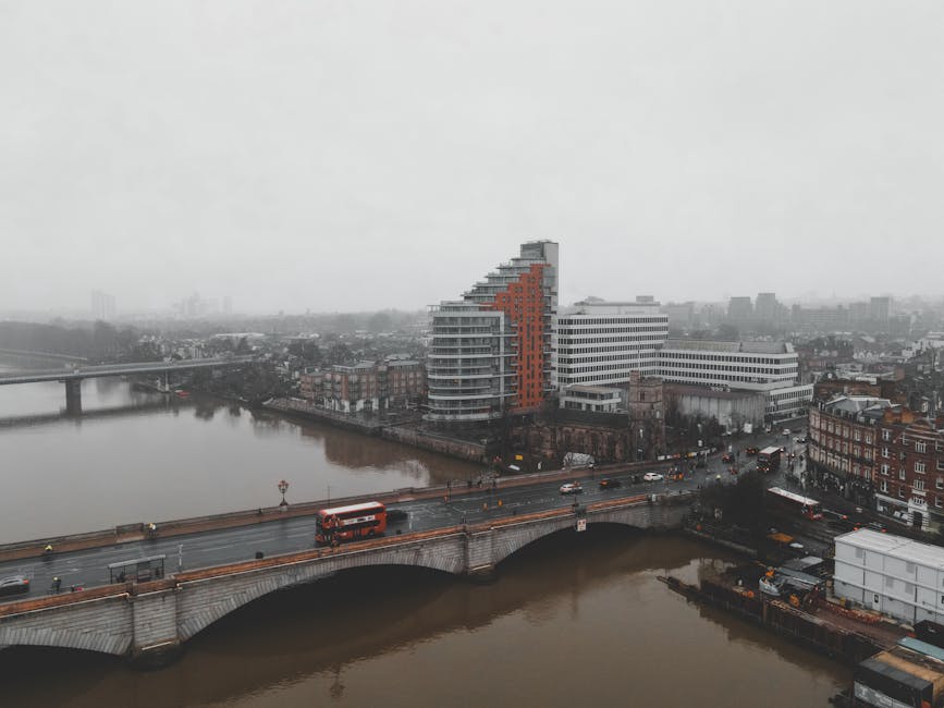 Aerial view of a cityscape on a cloudy day, featuring a modern, curved residential building with white and orange exterior panels situated along a riverbank. A bridge with vehicles crossing spans the river in the foreground, with surrounding traditional brick buildings and streets visible below. The scene is damp and overcast, with diffuse lighting highlighting the surfaces of the buildings and wet roadways. The overall atmosphere appears quiet and subdued, representative of typical urban scenery. This image emphasizes urban architecture and city infrastructure, relevant to the topic of surface cleaning and maintenance associated with residential environments, as addressed by Putney Carpet Cleaning in the context of mould remediation for Putney Embankment flats, PUTNEY.