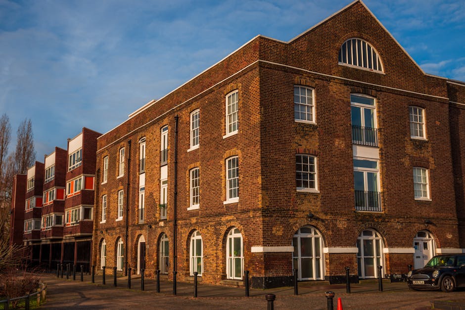 Exterior view of a multi-story residential brick building with large, white-framed windows and arched ground-floor entrances. The building is constructed from red and brown bricks, with some weathering visible on the facade. A clear blue sky with few clouds is above, and the sunlight casts warm tones on the surface. The area in front includes a paved pathway bordered by black metal posts and a parked car near the entrance. The surroundings are tidy and well-maintained, reflecting an urban residential environment. This image is related to surface cleaning and maintenance services provided by Putney Carpet Cleaning, emphasizing the importance of proper hygiene and deep cleaning for residential buildings like these.