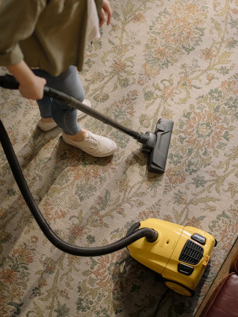 A person vacuuming a large, patterned area rug with a yellow and black canister vacuum cleaner in a room with wooden flooring partially covered by the rug. The individual, wearing beige shoes, light-colored jeans, and a beige jacket, is using the vacuum's hose attachment to clean the floral-designed carpet, which appears clean and well-maintained. The room is lit by natural or artificial light, highlighting the detailed pattern on the rug. The scene suggests a routine domestic surface cleaning process, emphasizing thorough deep cleaning and sanitisation by Putney Carpet Cleaning, a professional cleaning company. The focus is on the vacuuming activity within a living space, showcasing the equipment and the clean state of the carpet.