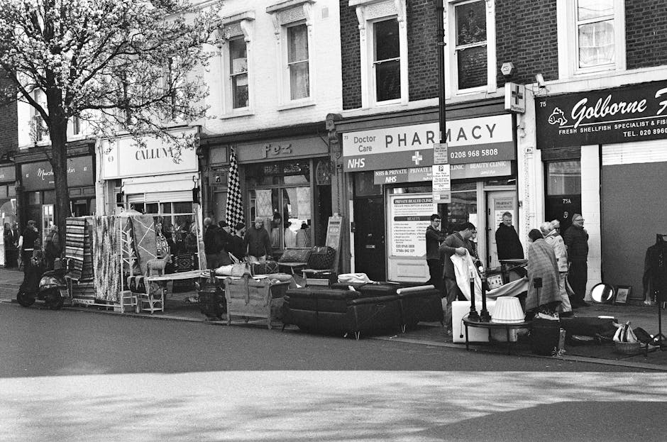 Street scene in Putney with a variety of second-hand furniture, including sofas and chairs, displayed on the sidewalk in front of shops. The shops have signs indicating a pharmacy and a fish and shellfish specialist, with a small crowd of people browsing and interacting. The scene is captured in black and white, featuring a mix of modern and older brick buildings, trees lining the street, and street furniture. The overall atmosphere suggests a local market or community event, with clear daylight illuminating the area, showing the outdoor displays and passersby. The image exemplifies a typical busy street scene in Putney, with an emphasis on community engagement and retail activity, relevant to urban living and local commerce.