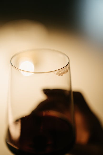 A close-up of a clear glass of red wine on a dark, reflective surface in a kitchen setting, with a blurred background emitting soft, warm lighting. The surface appears clean and free of residue, with the wine's deep color contrasting against the transparent glass. The lighting highlights the smooth curves of the glass and the rich texture of the wine, suggesting a tidy and well-maintained environment. This image relates to surface cleaning and maintenance, as featured in Putney Carpet Cleaning's emergency services for removing spills such as red wine stains in kitchens, demonstrating the importance of prompt deep cleaning and sanitisation in domestic spaces.