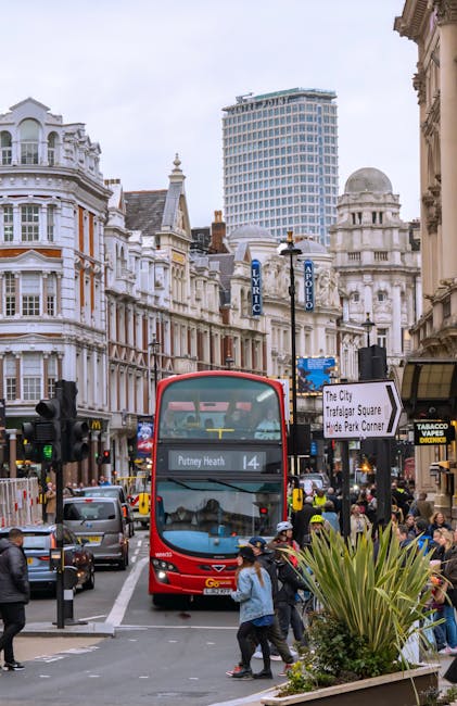 There is a busy street scene in Putney showing a double-decker red bus navigating through traffic, with pedestrians crossing the road and some waiting at the sidewalk. The buildings feature classic Victorian architecture with ornate white facades, large windows, and decorative detailing, set against a modern high-rise building in the background. The street is lined with street lamps, signs including a directional sign to Trafalgar Square and Hyde Park Corner, and storefronts such as a tobacco shop. The pavement appears clean and dry, and the scene is well-lit with natural daylight, reflecting a typical urban setting in Putney. This image relates to the environment where Putney Carpet Cleaning offers surface cleaning and sanitisation of residential and commercial spaces, ensuring hygiene and cleanliness in busy city areas.