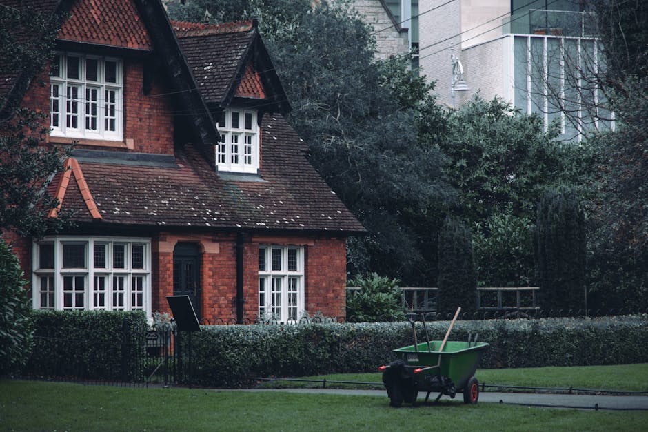A residential suburban house with red brick walls and a pitched roof covered with dark tiles, situated behind a well-maintained lawn and hedge. The house features multiple white framed windows, including a bay window on the ground floor and dormer windows on the upper level, all appearing clean and tidy. In the front yard, there is a green garden cart filled with tools such as a broom and a rake, indicating ongoing outdoor maintenance. Soft natural daylight illuminates the scene, highlighting the neatness of the garden and exterior surfaces, consistent with professional outdoor cleaning and maintenance standards. Putney Carpet Cleaning offers related services to maintain the appearance and hygiene of both indoor and outdoor surfaces.
