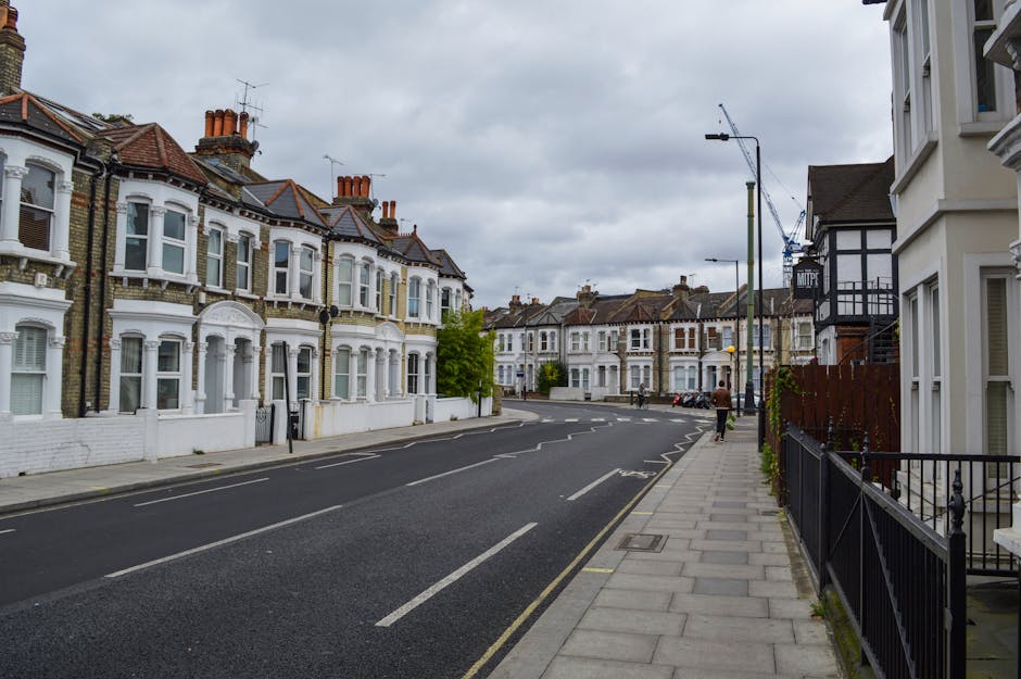 Image of a residential street in Putney, featuring a row of Victorian-style terraced houses with white facades, large bay windows, and red-tiled roofs. The street appears clean and well-maintained, with a paved sidewalk on the right side bordered by black metal railings and a few pedestrians walking along it. Overcast skies cast diffuse light over the scene. This setting highlights the type of environment where domestic cleaning services by Putney Carpet Cleaning are applicable, emphasizing the importance of surface cleaning, facade maintenance, and overall hygiene. Visible elements include asphalt road with clearly marked lanes, modern street lamps, and minimal vehicular presence, indicating a quiet neighbourhood likely benefiting from regular deep cleaning and sanitisation practices to maintain its tidy appearance.