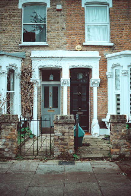 A residential suburban house with red brick walls and a pitched roof covered with dark tiles, situated behind a well-maintained lawn and hedge. The house features multiple white framed windows, including a bay window on the ground floor and dormer windows on the upper level, all appearing clean and tidy. In the front yard, there is a green garden cart filled with tools such as a broom and a rake, indicating ongoing outdoor maintenance. Soft natural daylight illuminates the scene, highlighting the neatness of the garden and exterior surfaces, consistent with professional outdoor cleaning and maintenance standards. Putney Carpet Cleaning offers related services to maintain the appearance and hygiene of both indoor and outdoor surfaces.